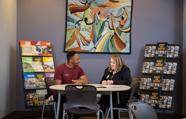 Two people are seated at a table in a room with informational brochures and a vibrant mural on the wall. 